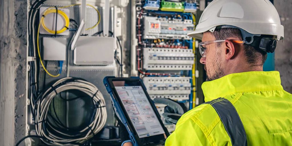 An electrician in safety gear uses a tablet to check a complex breaker panel during a routine electrical safety inspection.